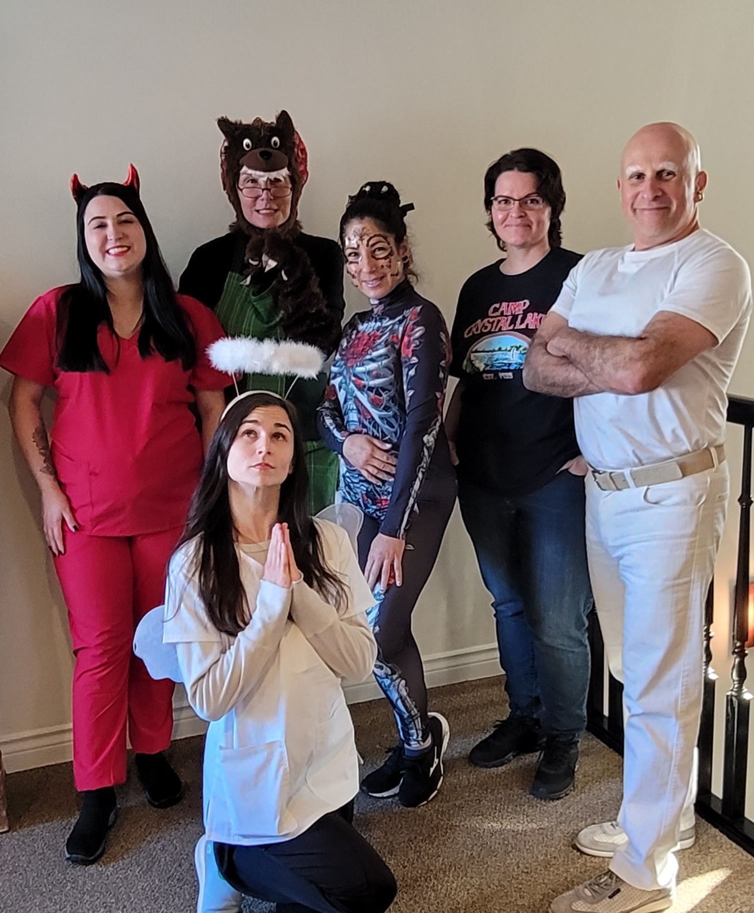 A group of six people in various costumes, including a devil, angel, and wolf, pose indoors in a casual setting.