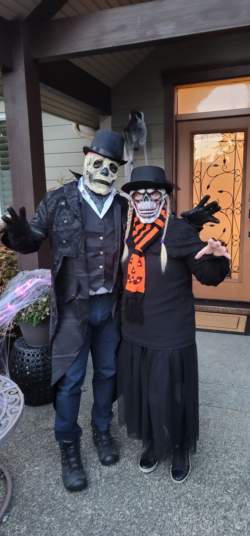 Two people in spooky skeleton masks, top hats, and dark formal attire pose together on a porch for Halloween.