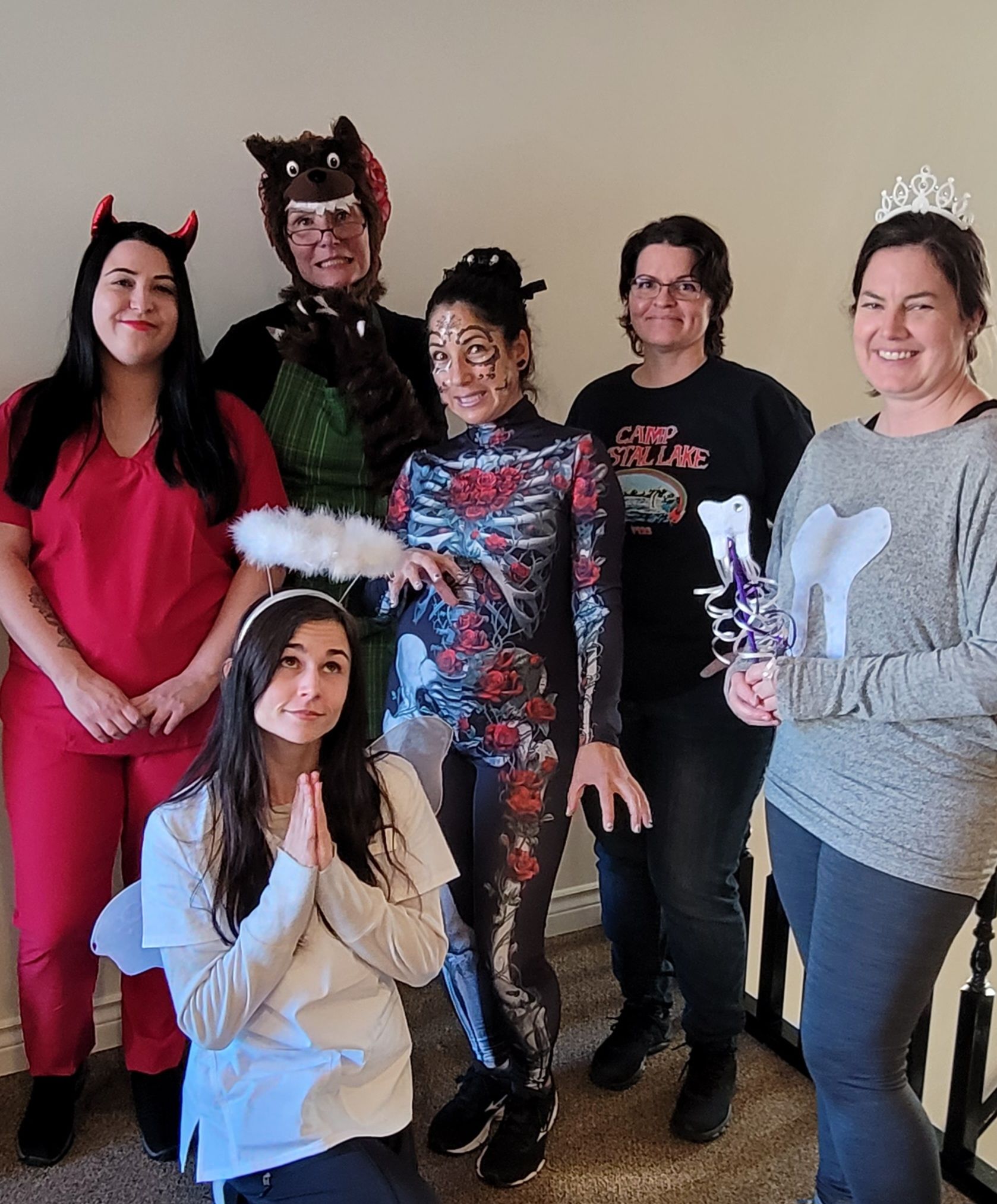 Six people pose in varied Halloween costumes, including a devil, a werewolf, an angel, a skeleton, and a tooth character.