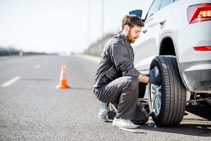 A technician in grey workwear kneels on a highway shoulder, changing a flat tire on a white SUV parked near a traffic cone.