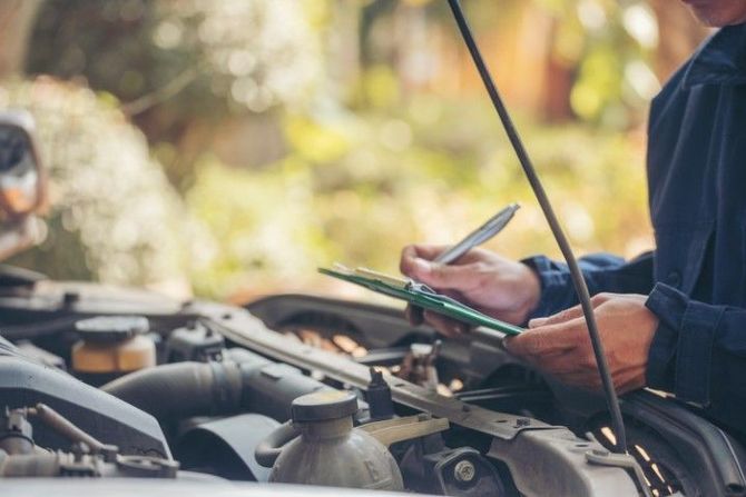 A mechanic writes on a clipboard while inspecting an open car engine, standing outdoors in daylight.