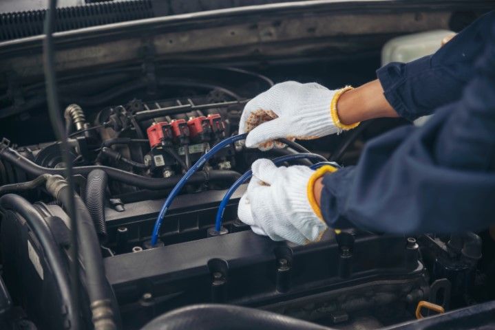 A mechanic wearing white gloves works on a car engine, holding blue ignition wires.