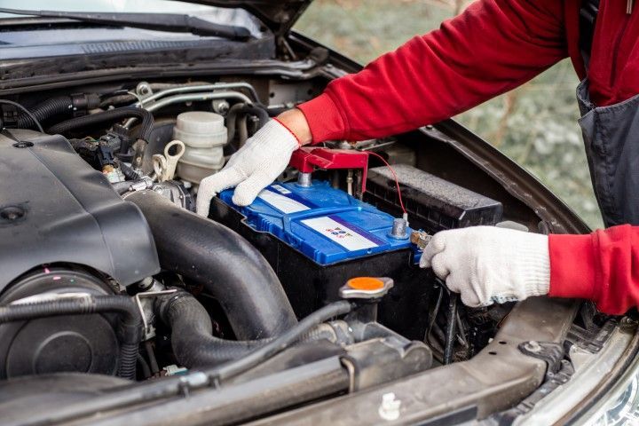 A mechanic in a red sweatshirt and white gloves installs a blue car battery into an open engine bay.
