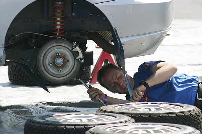 A mechanic in a blue shirt works on the rear axle and suspension of a white car elevated on a red jack stand.