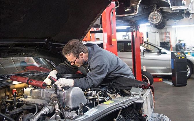 A mechanic in a grey hoodie works on an open car engine in a professional garage, with another car on a lift nearby.