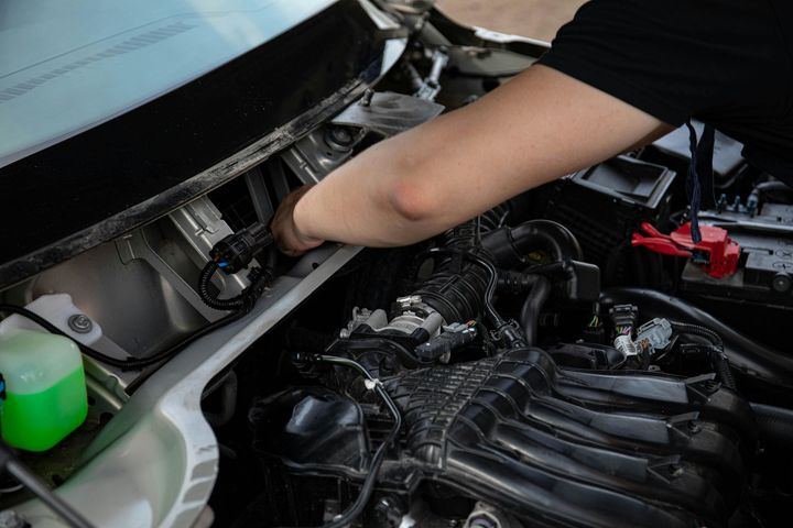 A person's arm reaches into the engine bay of a car to perform maintenance on wiring near the windshield.