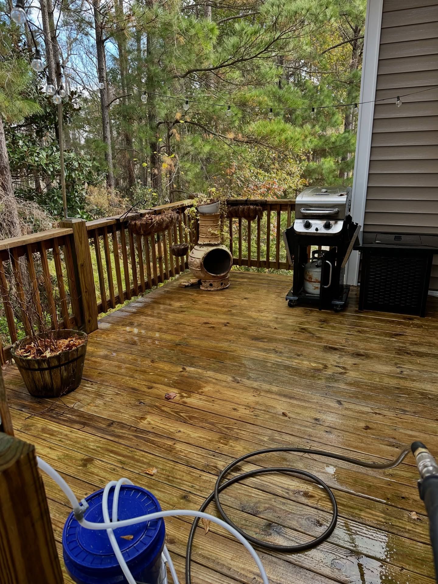 Wooden deck with grill, blue bucket, hose, and railing overlooking trees in a wooded yard.