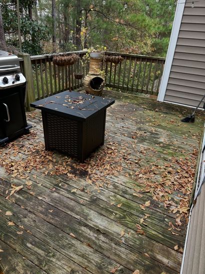 Wooden deck with a black fire pit table, grill, and a railing backed by trees and fallen leaves.