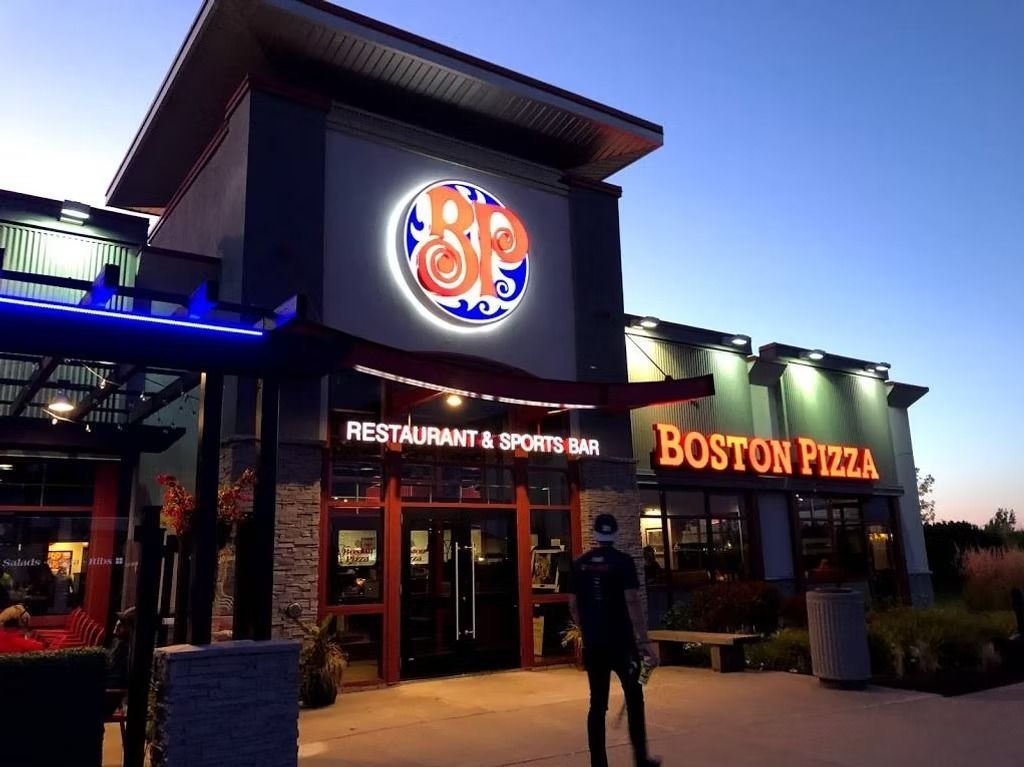 Boston Pizza restaurant exterior at dusk, neon logo, entrance, man standing.