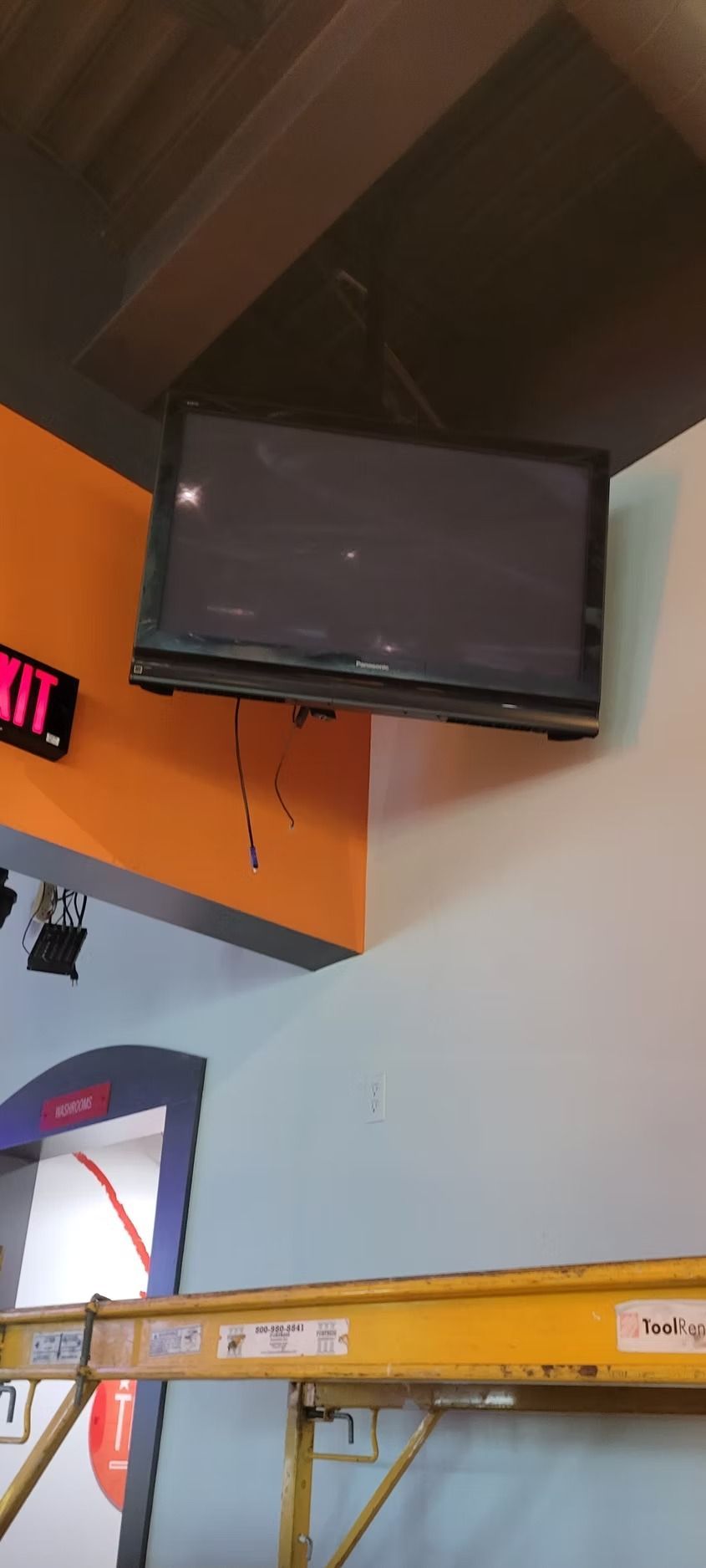 A TV mounted on a wall corner. Orange and white walls, with a yellow scaffold below.