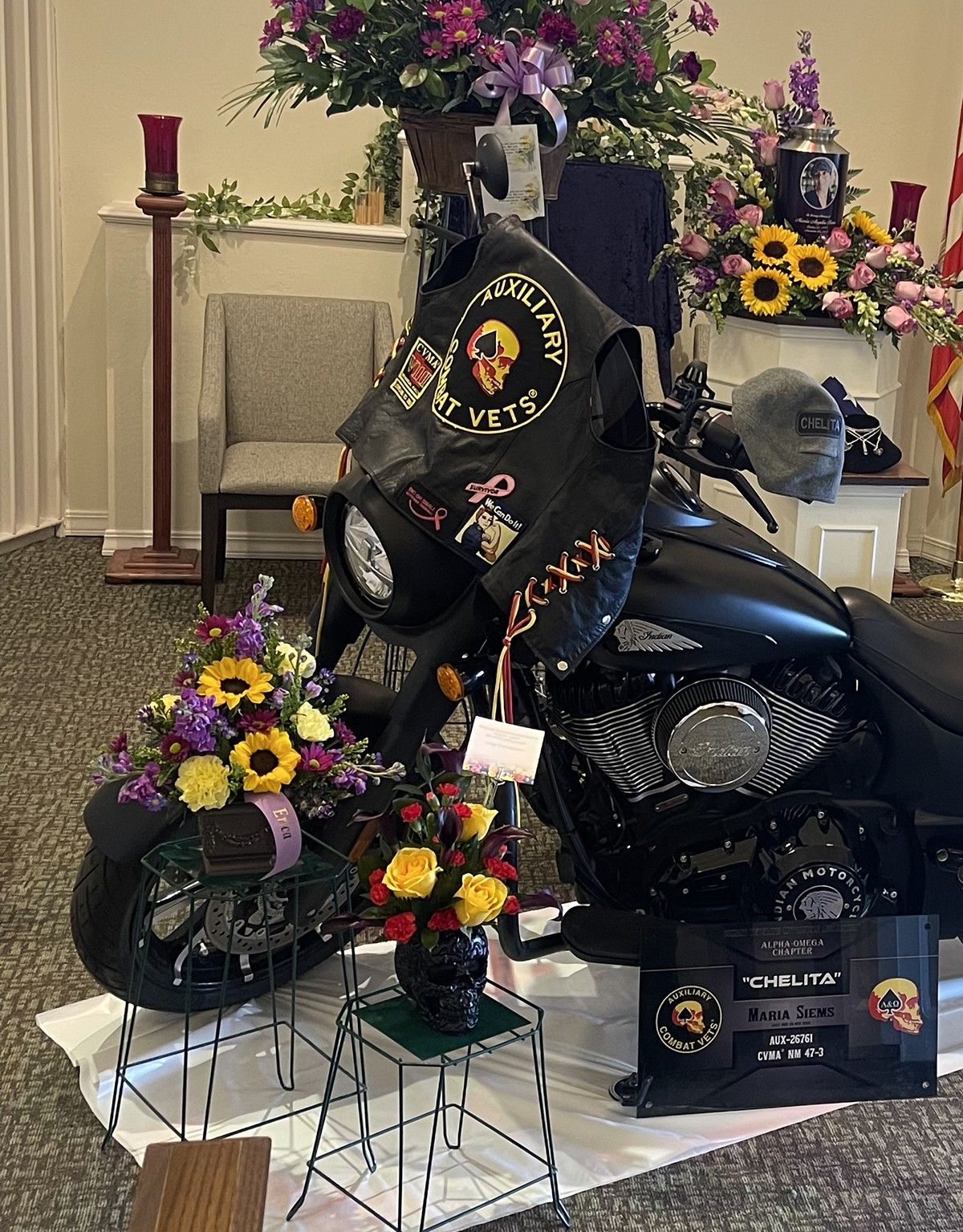 Memorial service display with motorcycle, rider vest, skull vase, CVMA tribute placard, and floral arrangements in a cremation tribute