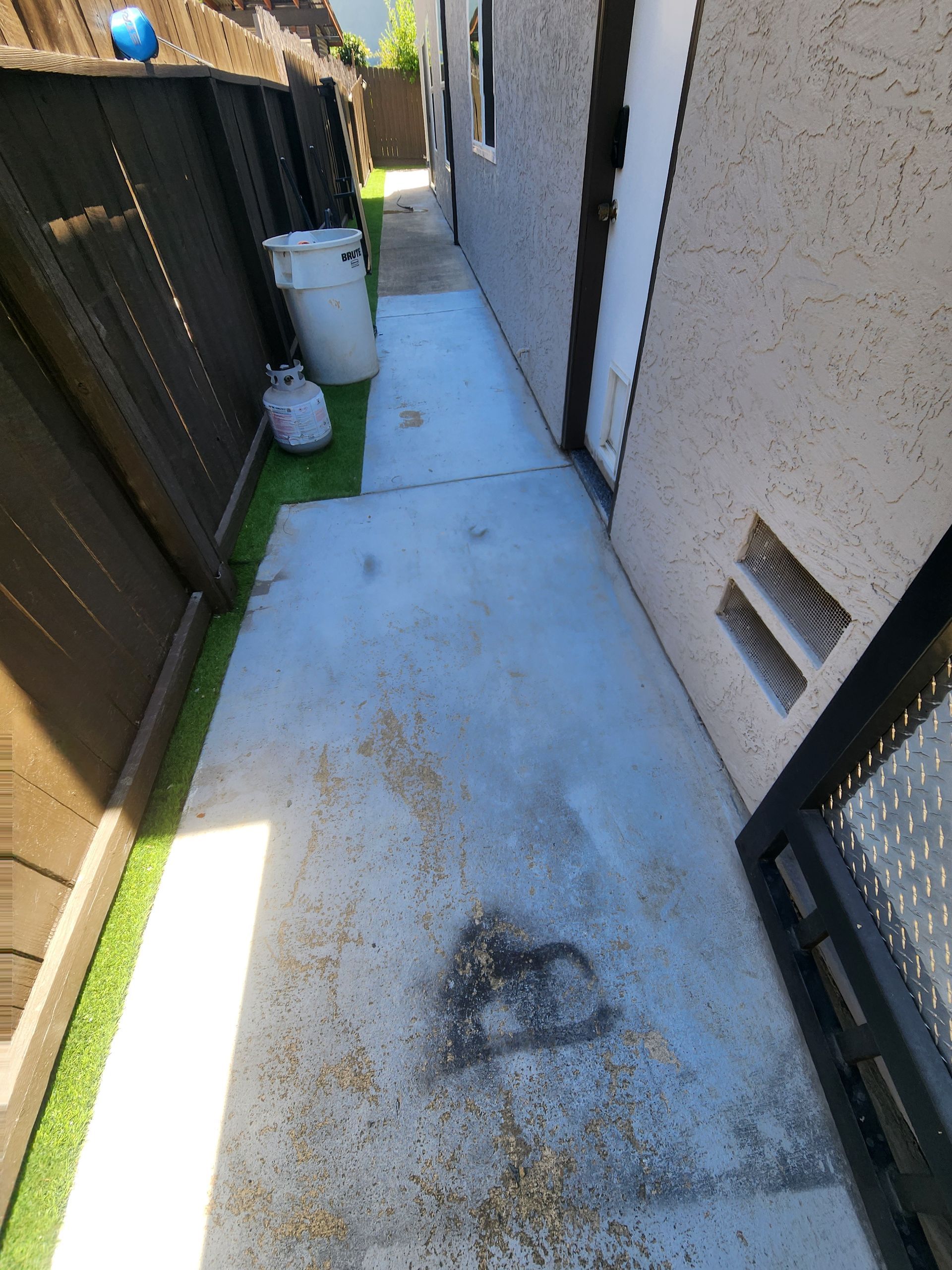 Narrow concrete side yard between a fence and house, with a small plant pot and vents on the wall.