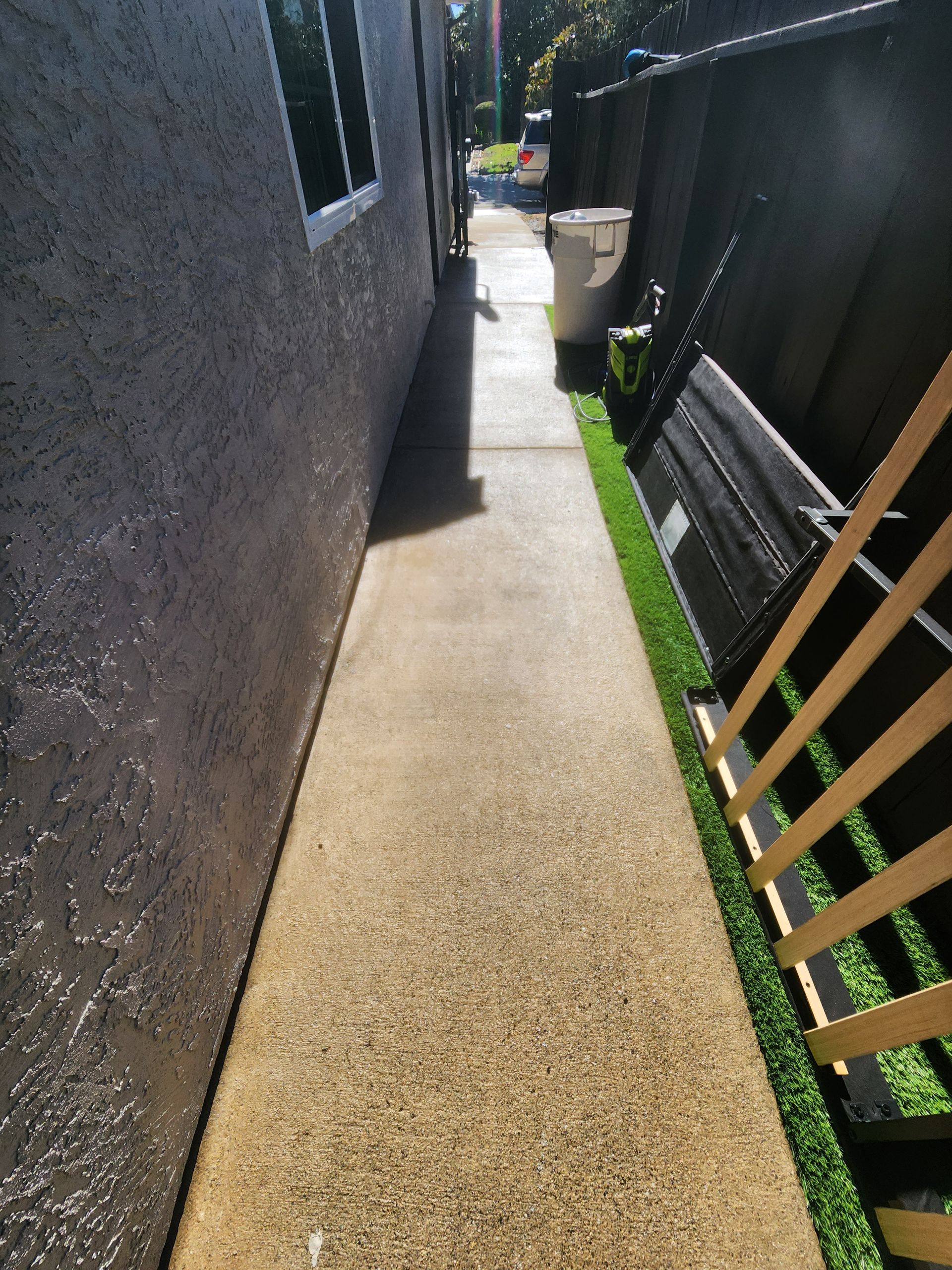 Narrow sunlit concrete walkway between a stucco wall and black fence, with a small wooden gate on the right.