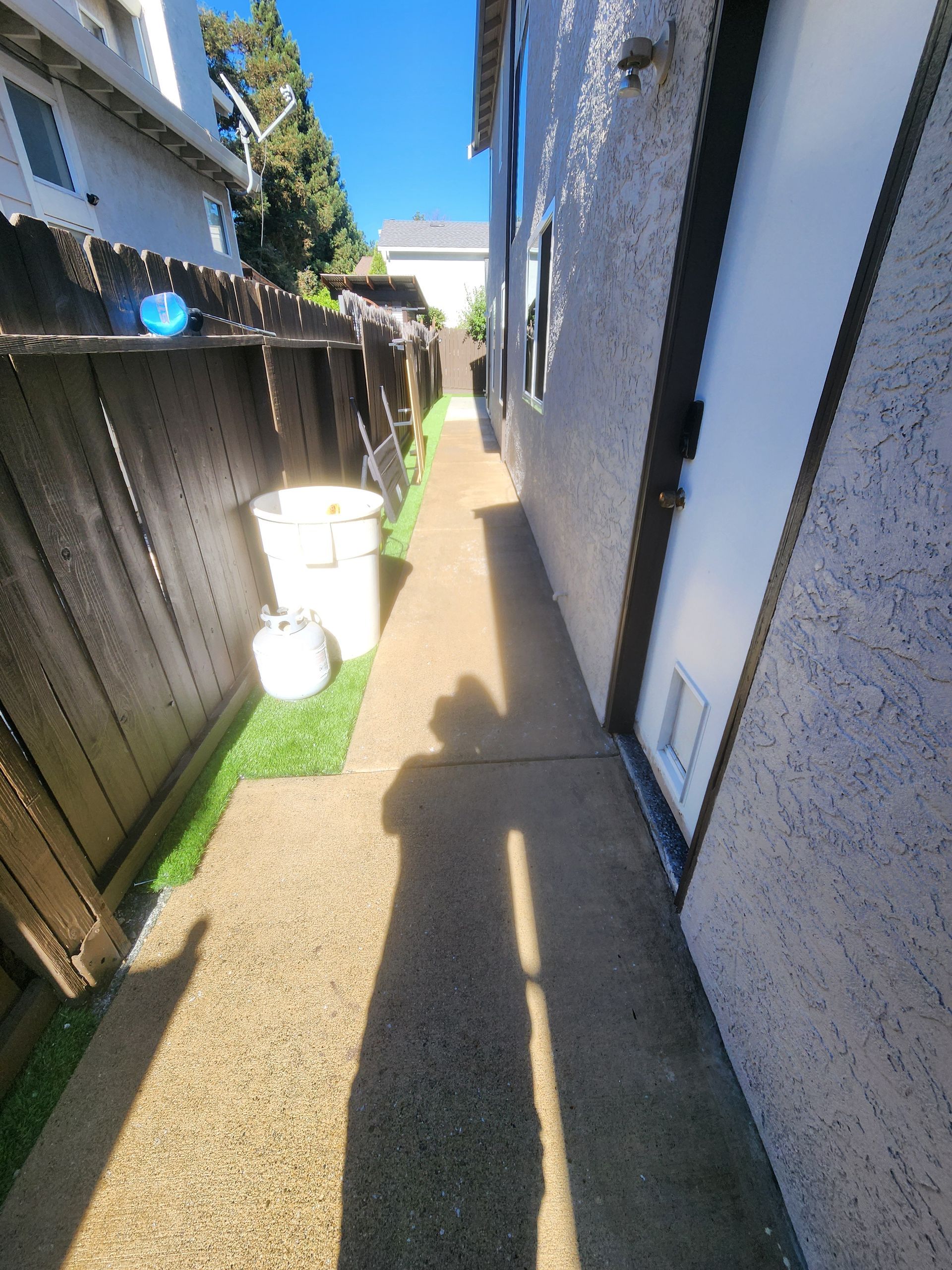Narrow outdoor side yard between house and fence, with white bins and sunlight casting long shadows.