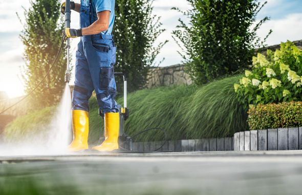 Person pressure washing a driveway beside green shrubs and a flowerbed, wearing yellow boots