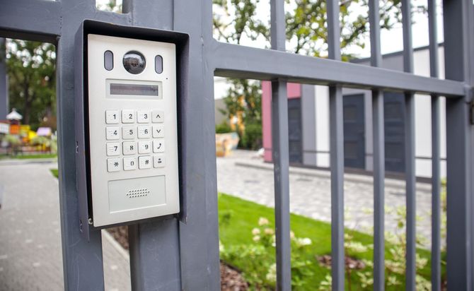 Silver intercom keypad mounted on a gray metal fence beside a building entrance