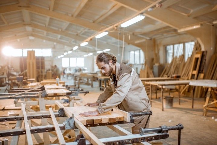 Woodworker sanding a plank in a bright workshop with stacked lumber and workbenches
