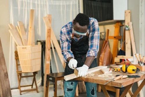 Person in a workshop cutting wood at a workbench, wearing gloves and a face mask.