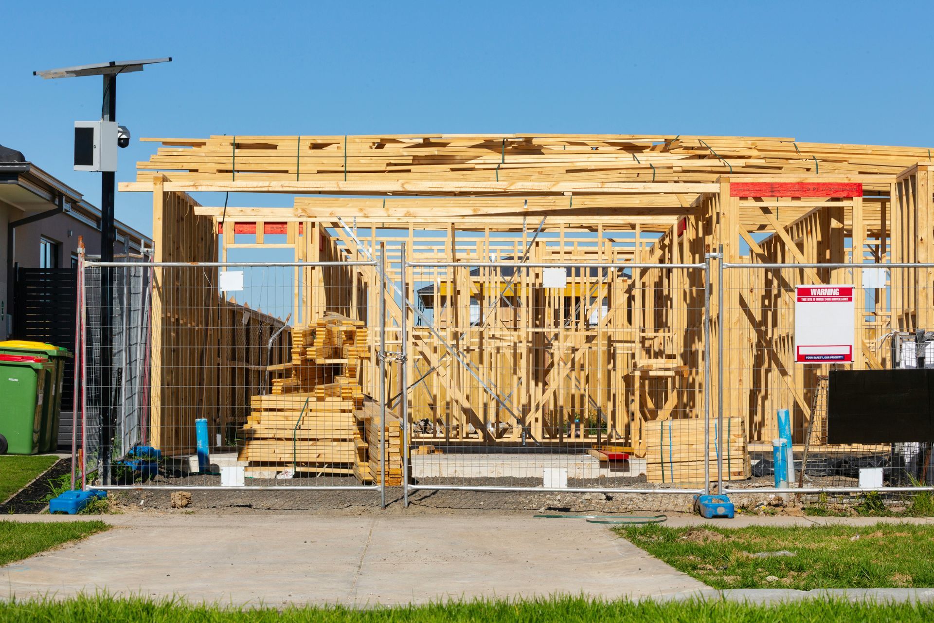 Wood-framed house under construction with exposed beams, scaffolding, and tools in a fenced lot