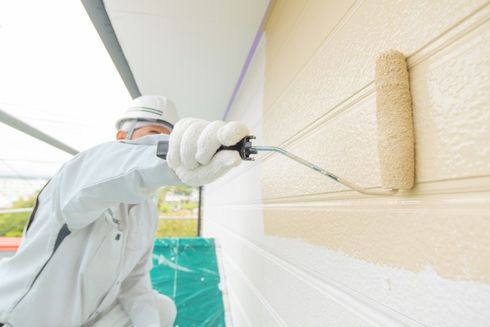 Painter in white coveralls rolling beige paint onto an exterior wall under a covered walkway