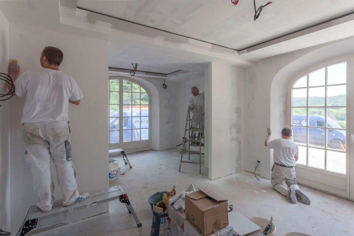 Painters in white overalls working in a bright, unfinished room with large arched windows.
