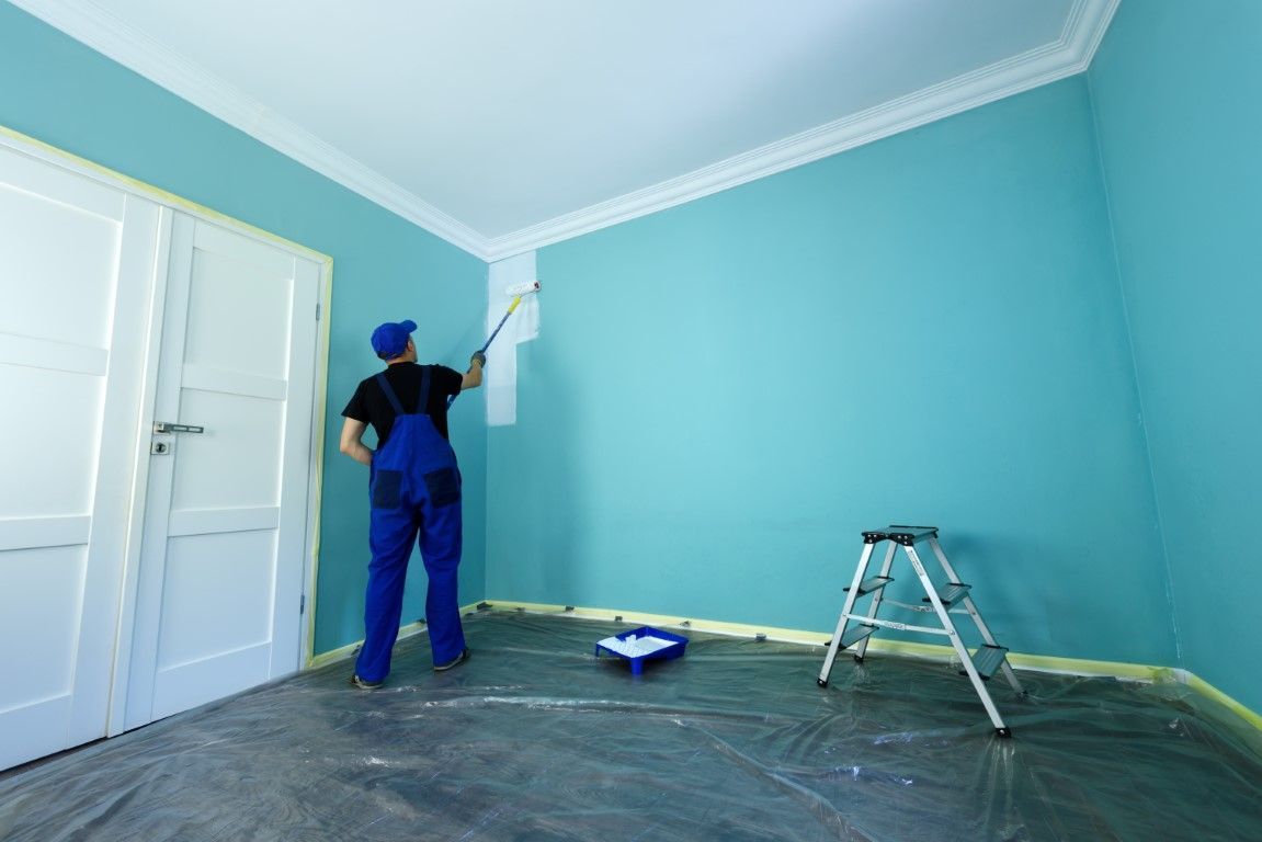 Painter in blue overalls painting a turquoise room near a white door, with a ladder and paint tray on the floor