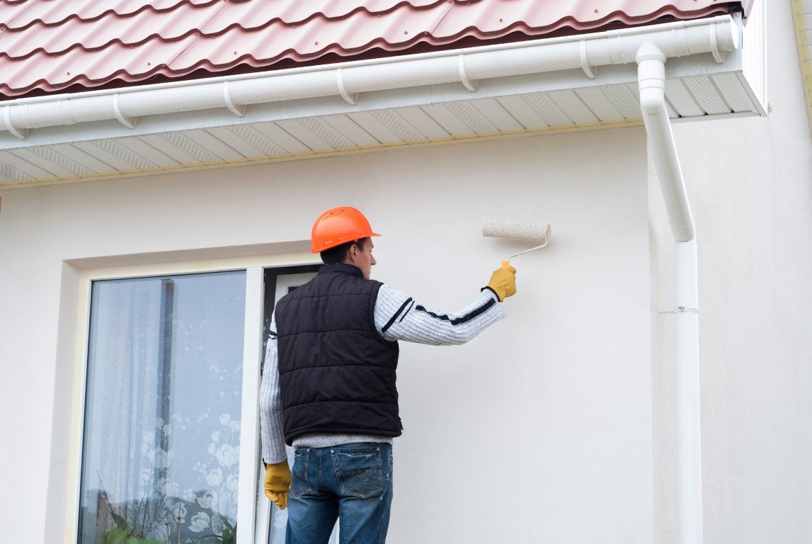 Worker painting a white house exterior under the roofline, wearing an orange hard hat and black vest
