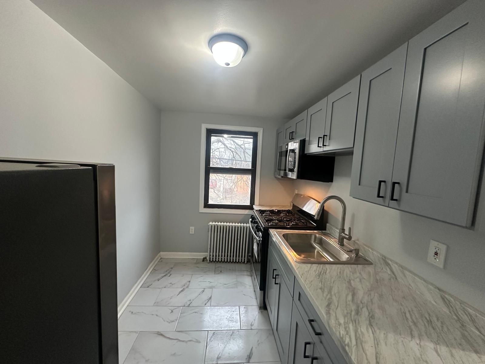 A small kitchen with gray cabinets, white countertops, and a black stove, a window, and a refrigerator.