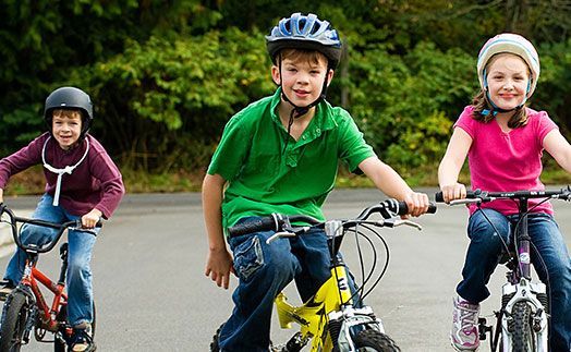 Three children wearing helmets ride bicycles on a paved road outdoors, smiling toward the camera.