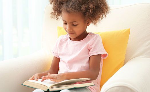 A person with curly hair wearing a pink shirt sits in a white armchair and reads an open book in front of a yellow pillow.