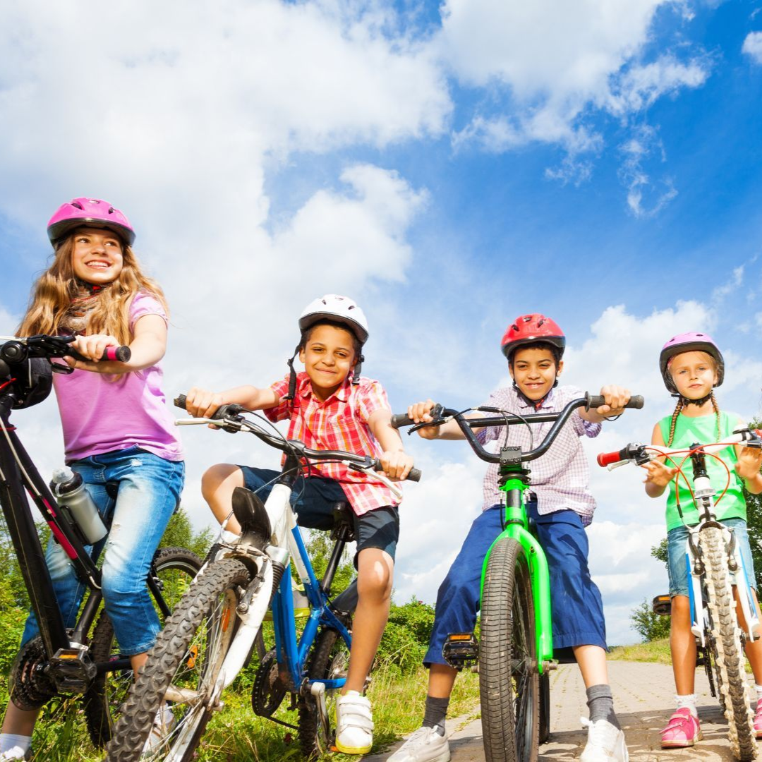 Four smiling children in helmets ride their bicycles on a path under a bright blue, sunny sky.