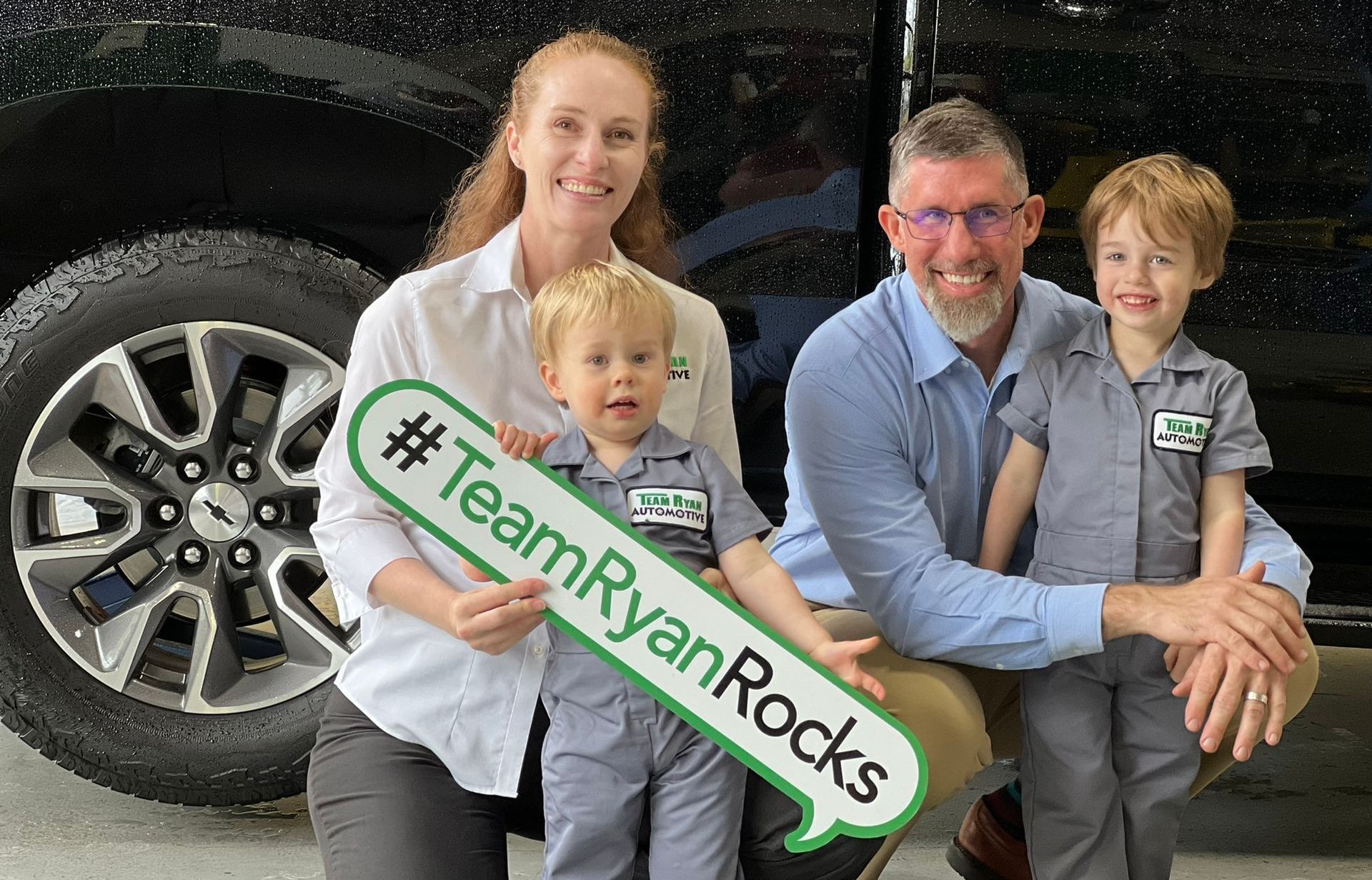 Family poses with hashtag sign, smiling, near a vehicle.