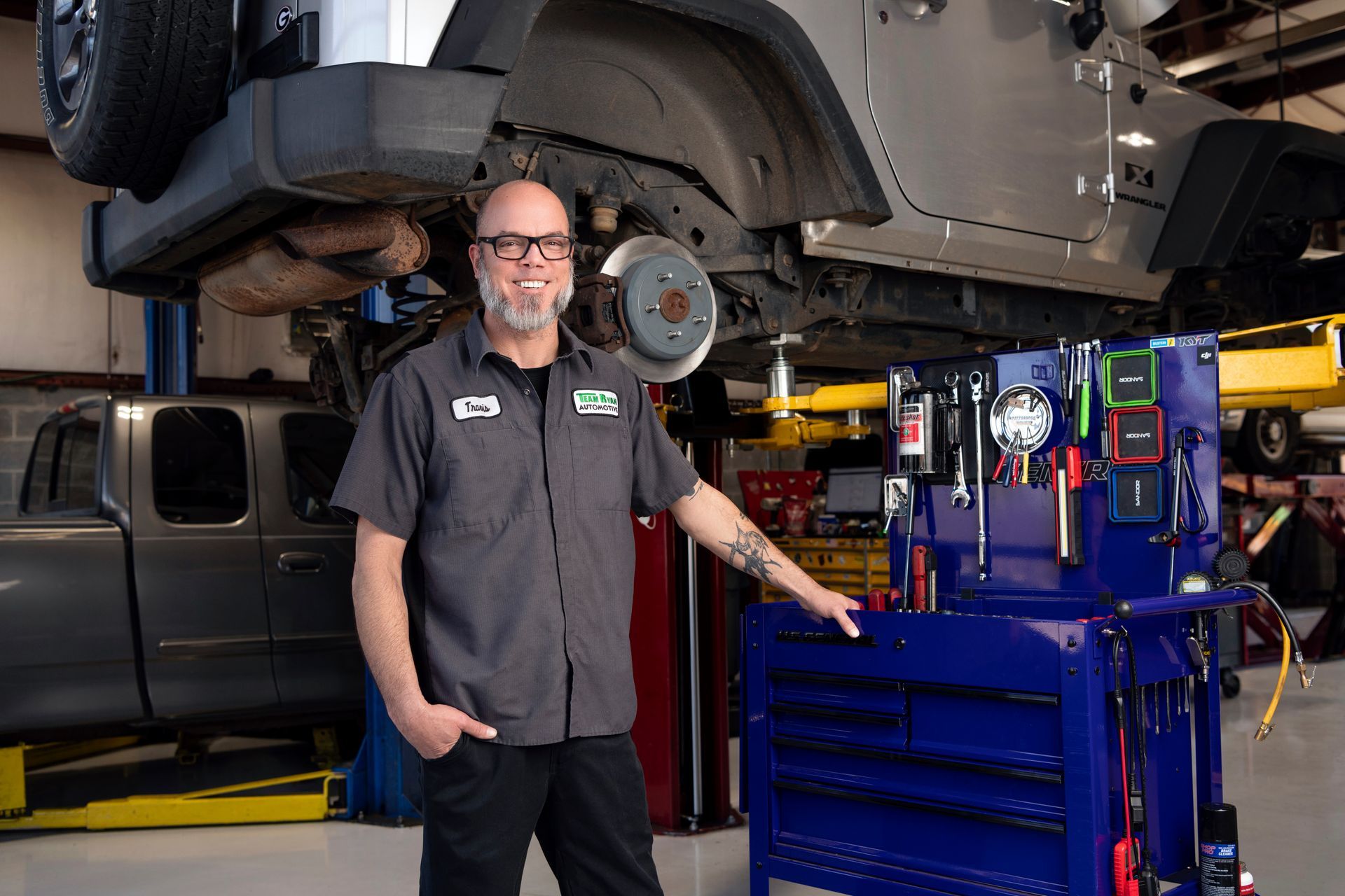 Mechanic standing beside a car lifted on a hoist in an auto repair garage, with tools nearby