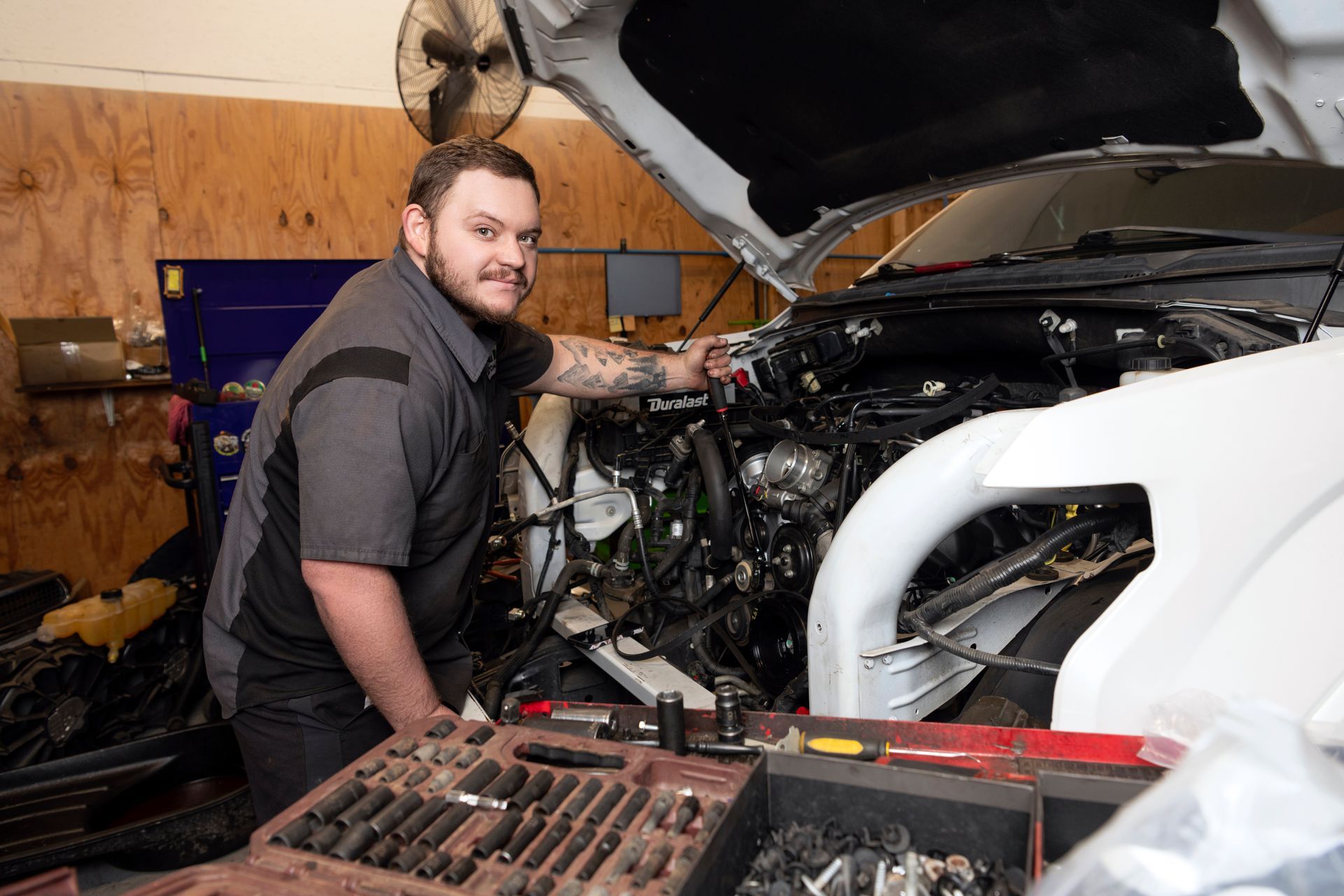 Mechanic working on a white car with the hood open in a garage, tools in the foreground.