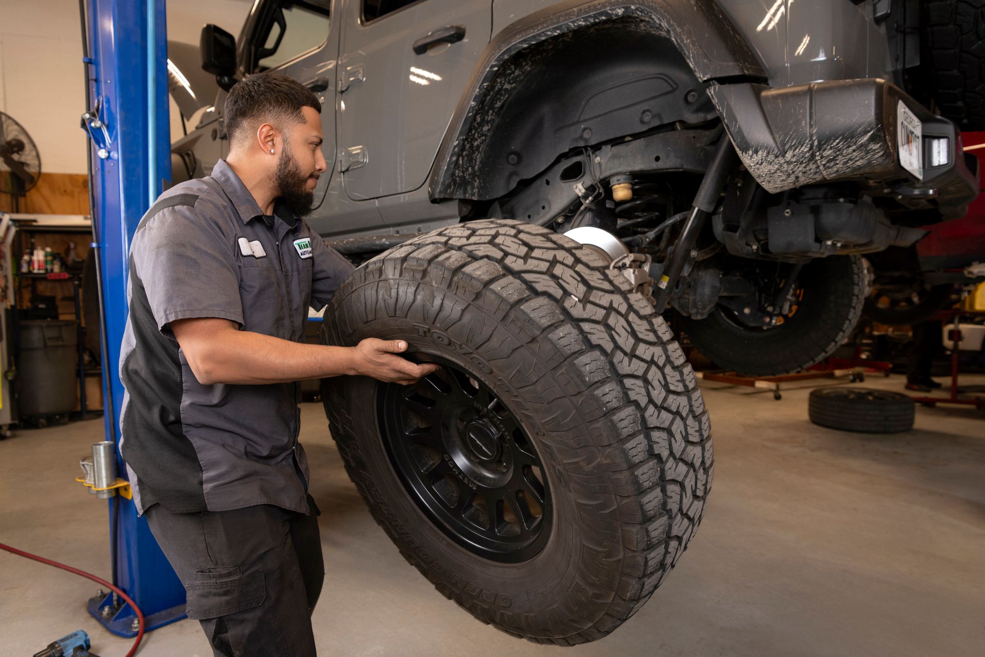 Mechanic lifting a large off-road tire beside a vehicle on a shop lift