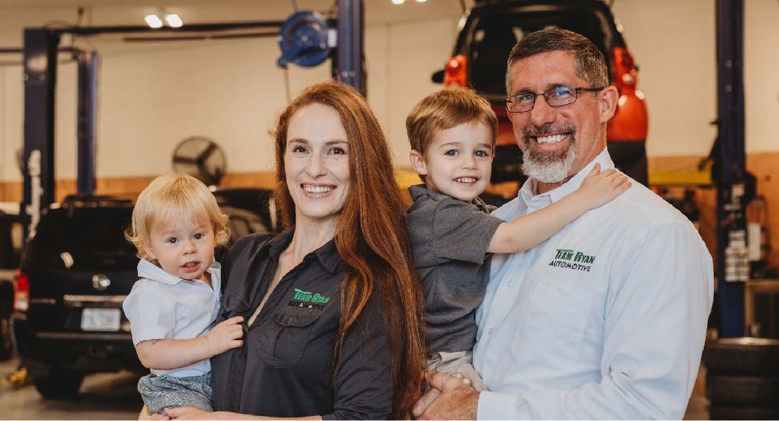 Family of four smiling in an auto shop, with a mechanic holding a child and garage equipment behind them