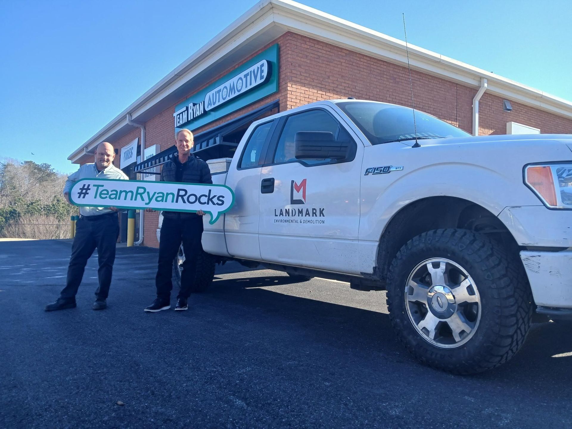 Two people holding #TeamRyanRocks sign beside a white WorkMax truck outside a brick building.