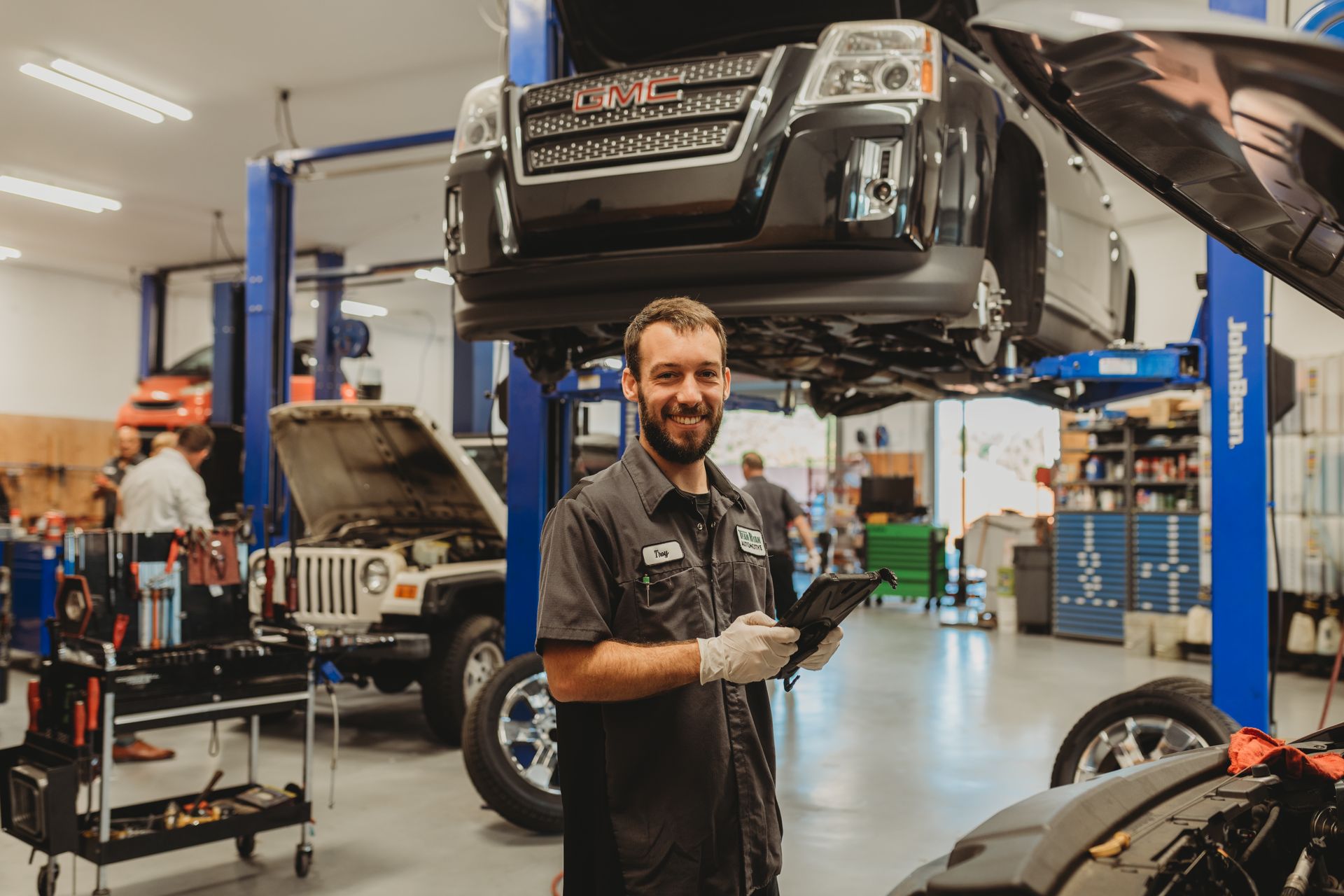 Mechanic smiling in an auto repair shop with a car lifted on a blue hoist.