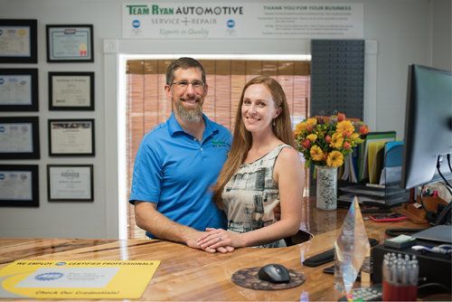 Two people standing behind a front desk in an automotive office, smiling with flowers and framed certificates nearby.