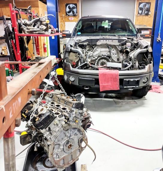 A truck with an open engine bay, new engine on a hoist in a garage.