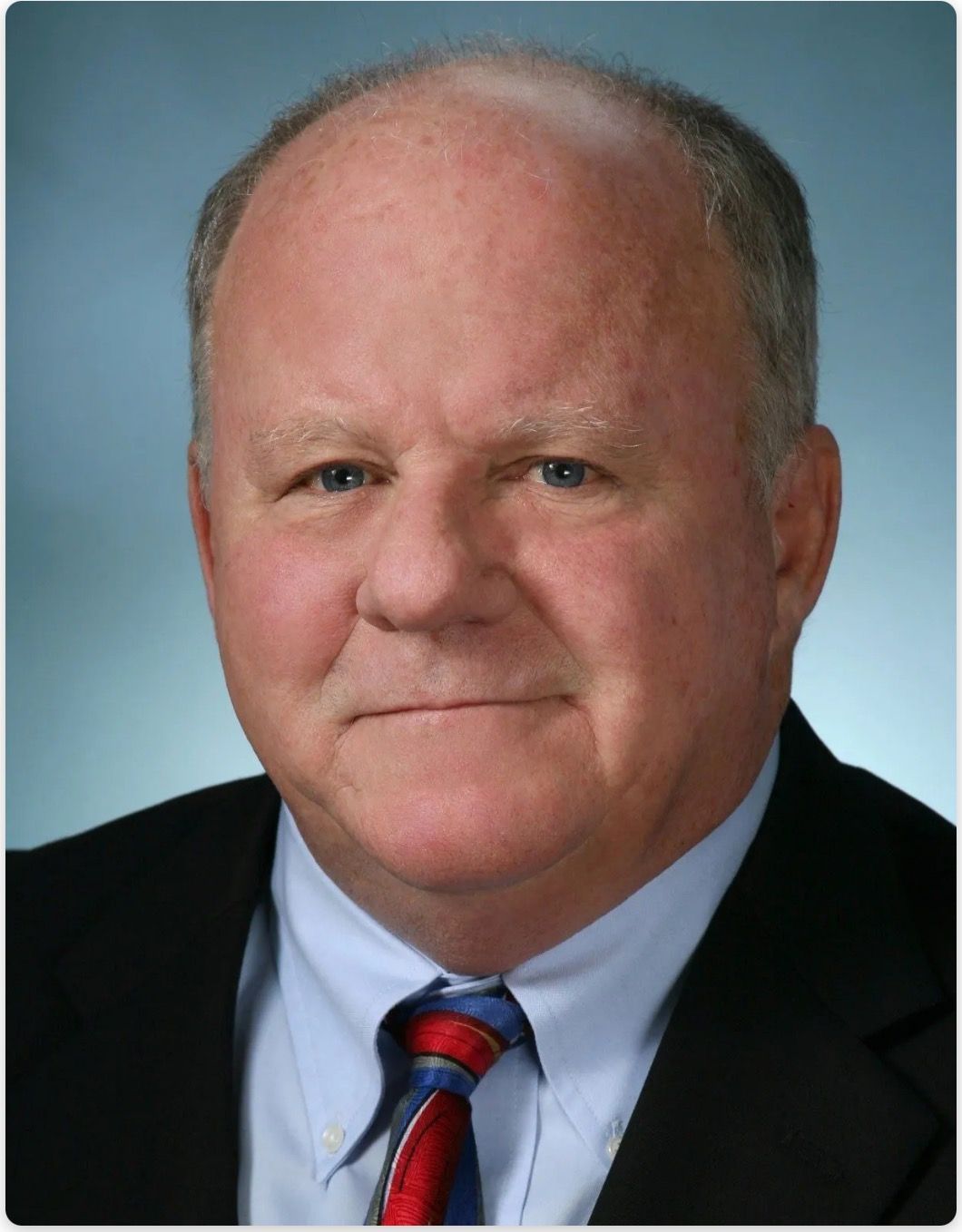 Studio headshot of a man in a dark suit, light blue shirt, and red striped tie against a blue background