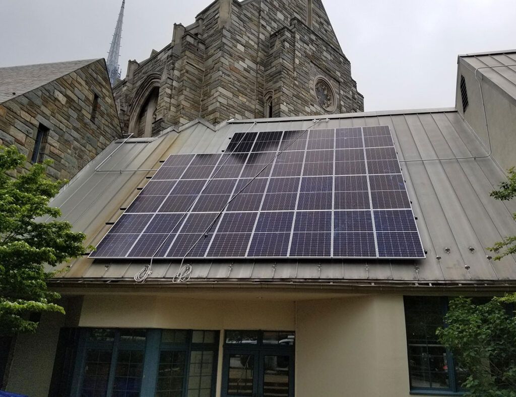 Building roof with solar panels under a cloudy sky, with a stone church tower in the background