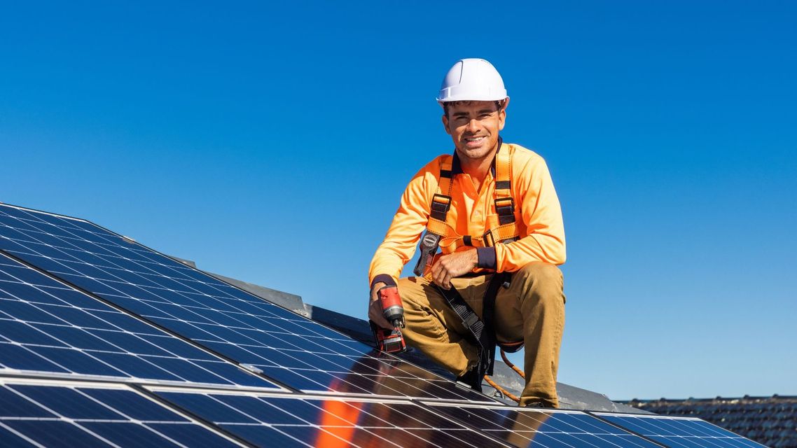 Worker in hard hat kneeling on rooftop solar panels under a clear blue sky