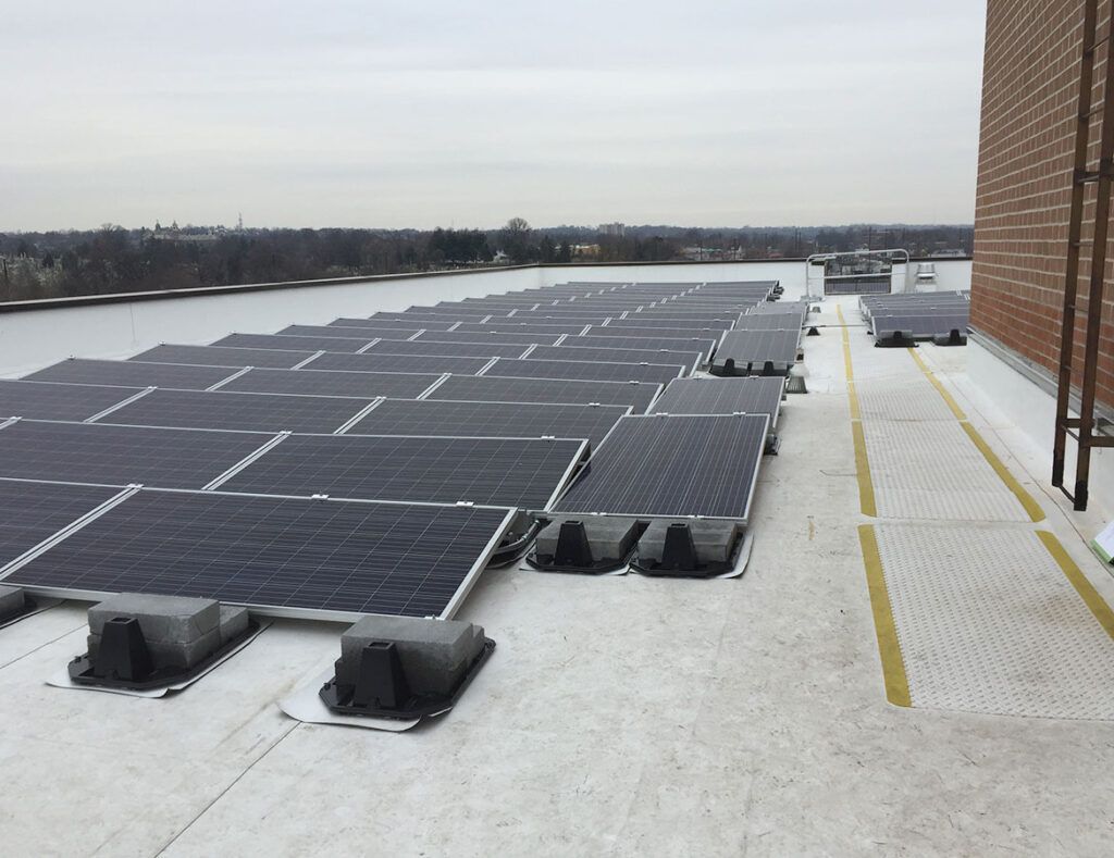 Snow-covered rooftop with rows of solar panels and a walkway beside a brick building.