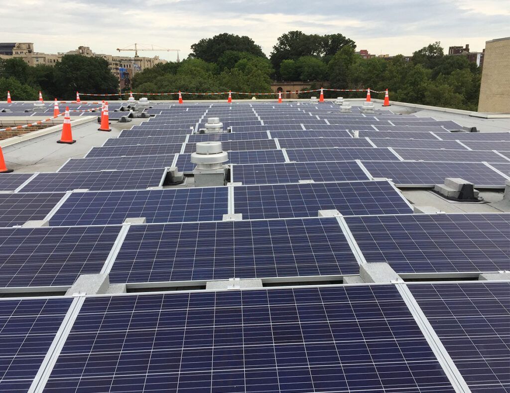 Rows of blue solar panels on a rooftop under an overcast sky, with orange safety cones in the distance