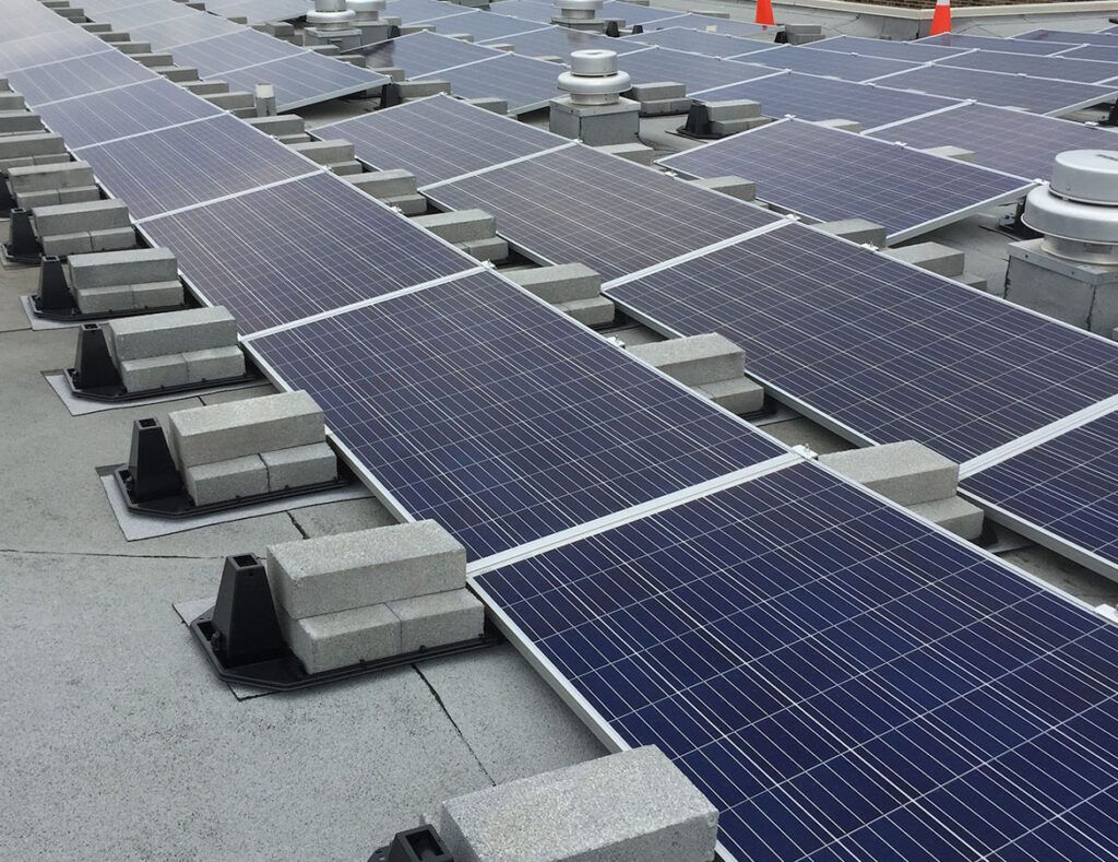 Rows of blue solar panels mounted on a rooftop with gray ballast blocks and orange cones in the background