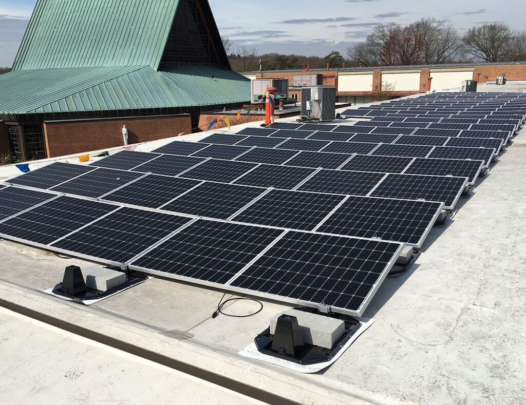 Rows of solar panels on a flat rooftop beside a green-roofed building under a partly cloudy sky