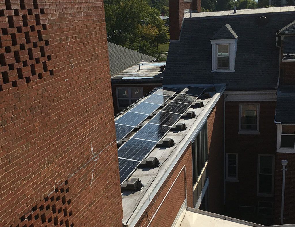 Rooftop solar panels beside a brick building and a dark-roofed house in an urban courtyard.