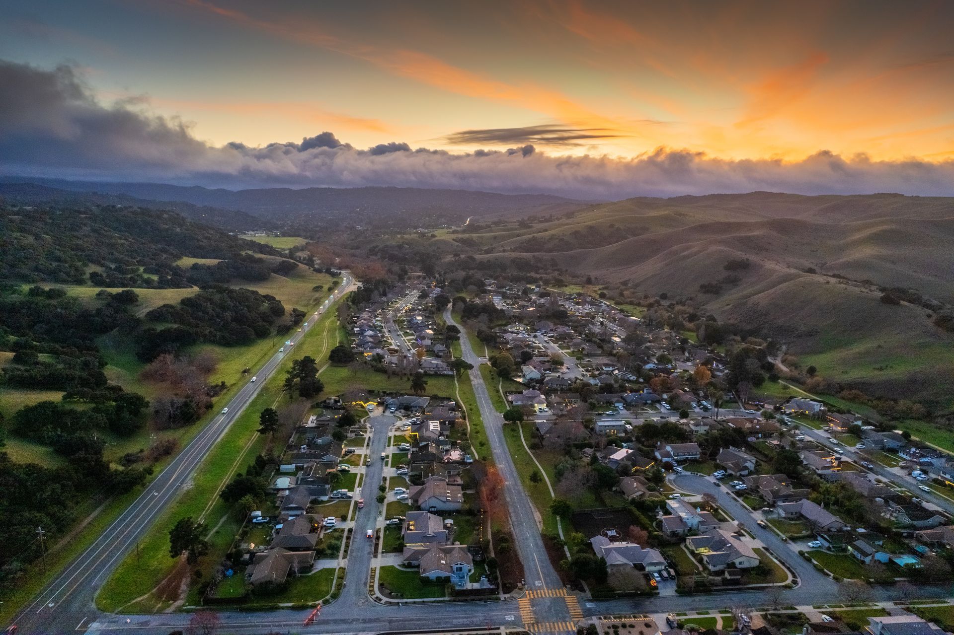 Aerial view of a suburban neighborhood at sunset with a highway on the left and hills in the background.