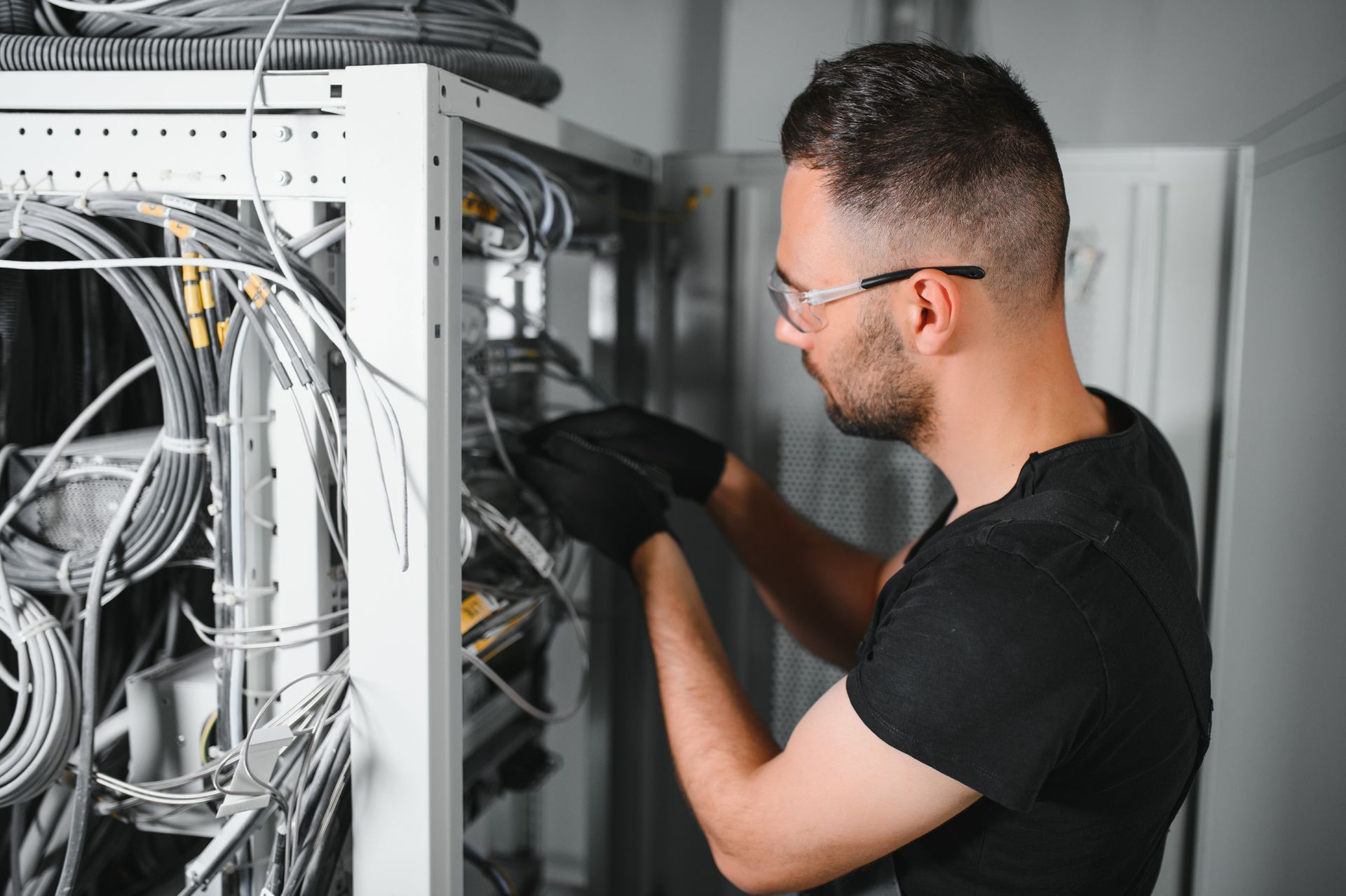 Man in black gloves and glasses working on a server rack filled with cables.
